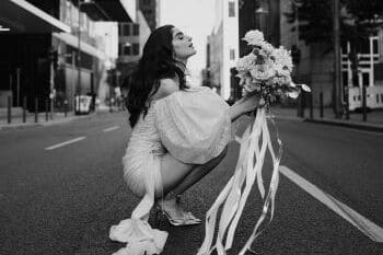 Braut hockend auf Straße mit Brautstrauß und kurzem Hochzeitskleid