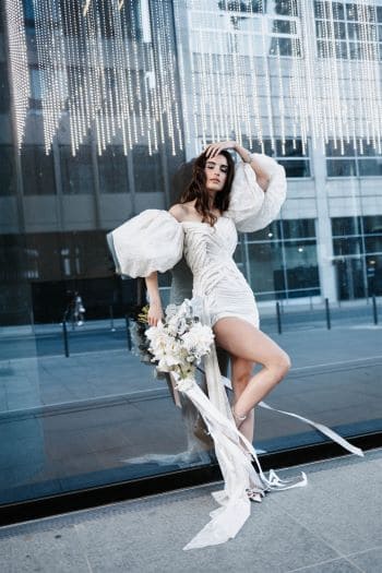 Braut lehnend an Glasfront mit kurzen Hochzeitskleid mit Puffärmeln